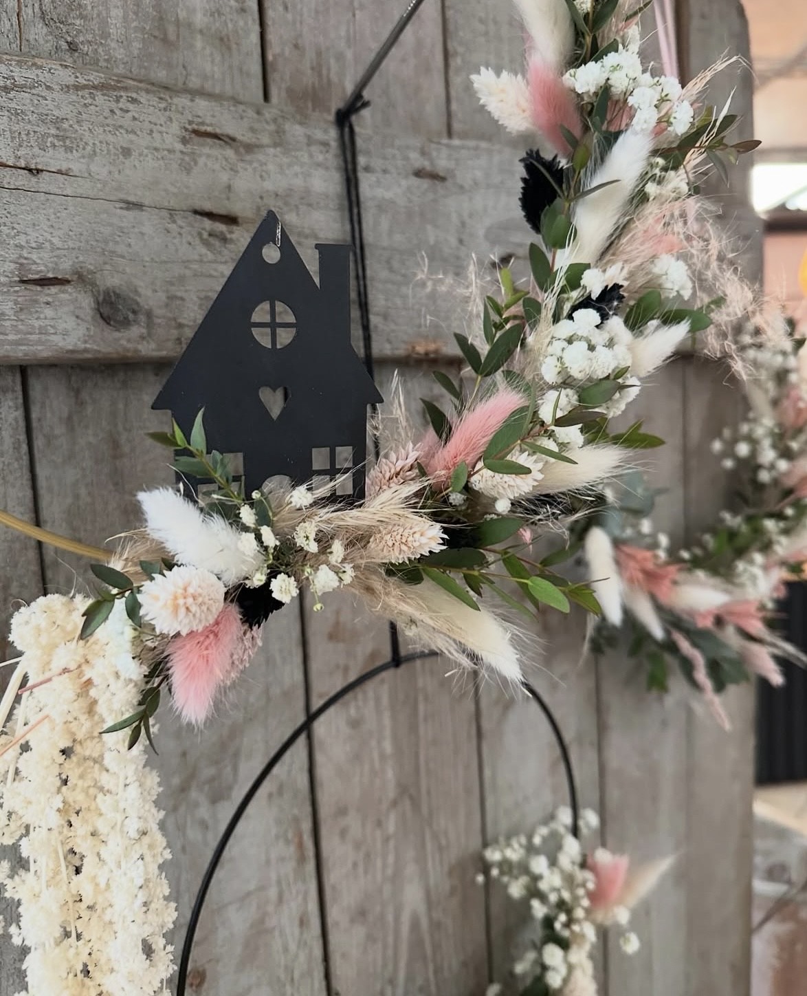 Ein filigraner Kranz aus weißen und rosa Trockenblumen mit grünem Laub und einem schwarzen Hausanhänger vor einer rustikalen Holzwand